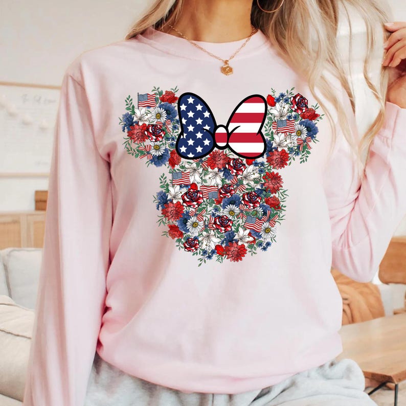 May include: A light pink long-sleeve sweatshirt featuring a floral design in the shape of Mickey Mouse ears. The design includes a bow with a stars and stripes pattern, and red, white, and blue flowers and American flags.