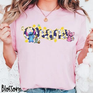 Puede incluir: Camiseta rosa claro con la palabra "Ohana" en un dise&ntilde;o floral azul con detalles amarillos. La camiseta presenta los personajes de dibujos animados Stitch y Angel, con elementos tem&aacute;ticos de Pascua. La palabra "Blossom" est&aacute; impresa en la parte inferior.