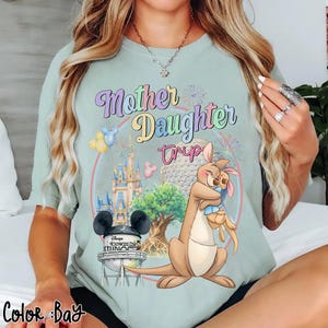 May include: A pale green t-shirt featuring the text "Mother Daughter Trip" in pastel hues. The design includes a kangaroo embracing a joey, a castle, and a water tower with Mickey Mouse ears. The words "Disney's Hollywood Studios" are also present.