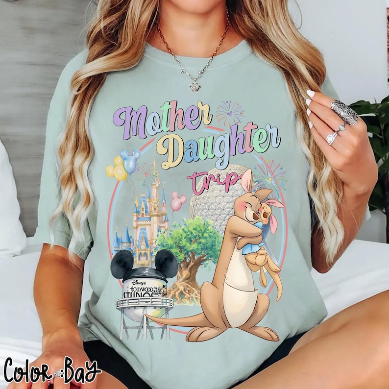 May include: A pale green t-shirt featuring the text "Mother Daughter Trip" in pastel hues. The design includes a kangaroo embracing a joey, a castle, and a water tower with Mickey Mouse ears. The words "Disney's Hollywood Studios" are also present.