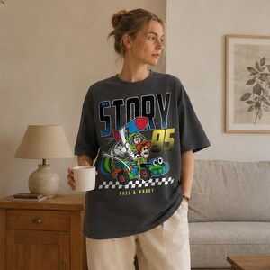 May include: Dark gray t-shirt featuring a colorful graphic of Buzz Lightyear and Woody from Toy Story in a race car with a rocket. The text "TOY STORY 95" and "BUZZ & WOODY" are also printed on the shirt.