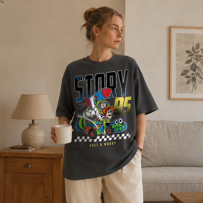 May include: Dark gray t-shirt featuring a colorful graphic of Buzz Lightyear and Woody from Toy Story in a race car with a rocket. The text "TOY STORY 95" and "BUZZ & WOODY" are also printed on the shirt.