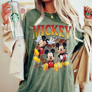 May include: An olive green t-shirt with the words "Mickey Mouse" in yellow and red, and three cartoon images of Mickey Mouse. The shirt has a crew neck and short sleeves.