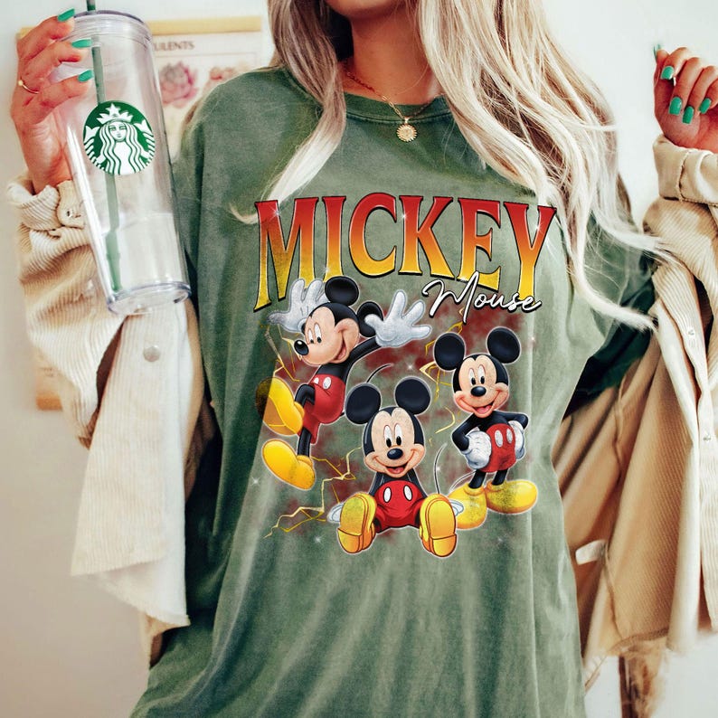 May include: An olive green t-shirt with the words "Mickey Mouse" in yellow and red, and three cartoon images of Mickey Mouse. The shirt has a crew neck and short sleeves.