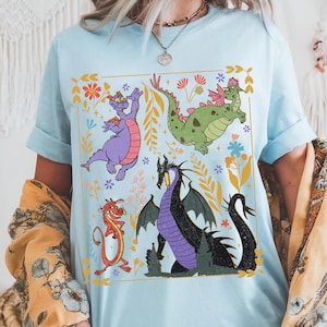 May include: Light blue t-shirt featuring a colorful graphic of cartoon dragons and floral elements. The design includes a purple dragon, a green dragon, a red dragon, and a black dragon. The shirt has short sleeves and a crew neck.