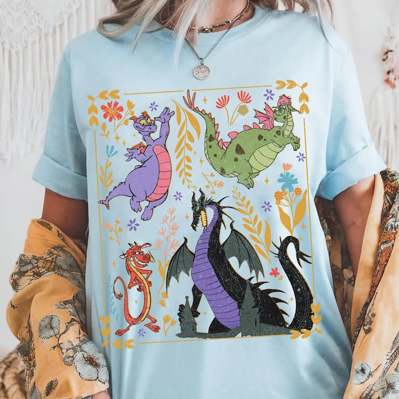May include: Light blue t-shirt featuring a colorful graphic of cartoon dragons and floral elements. The design includes a purple dragon, a green dragon, a red dragon, and a black dragon. The shirt has short sleeves and a crew neck.