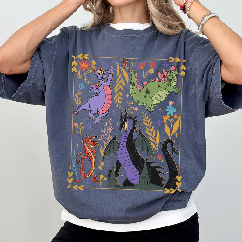 May include: A blue-grey t-shirt featuring a colorful illustration of dragons and floral elements within a gold rectangular frame. The design includes a purple dragon, a green dragon, a red dragon, and a black dragon. The shirt has short sleeves and a crew neck.