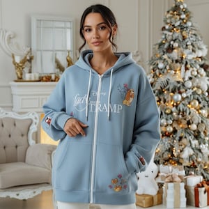 May include: A light blue zip-up hoodie featuring the text "Lady and the Tramp" with a graphic of the dog characters. The hoodie has a drawstring hood, front pockets, and embroidered floral and dog details on the sleeves and front.