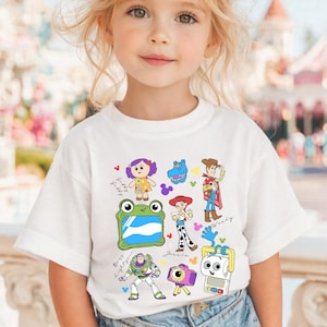 May include: White short-sleeved t-shirt featuring colorful cartoon characters from the Toy Story franchise. The design includes Woody, Buzz Lightyear, Jessie, and other characters. The shirt is worn by a young child.