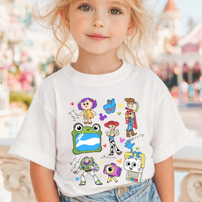 May include: White short-sleeved t-shirt featuring colorful cartoon characters from the Toy Story franchise. The design includes Woody, Buzz Lightyear, Jessie, and other characters. The shirt is worn by a young child.