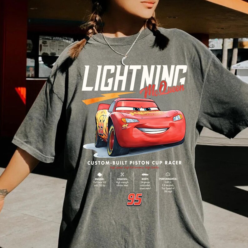 Puede incluir: Camiseta gris con Lightning McQueen de la pel&iacute;cula Cars. El dise&ntilde;o incluye el texto "LIGHTNING McQueen" sobre un coche de carreras rojo con el n&uacute;mero 95. El texto "CUSTOM-BUILT PISTON CUP RACER" est&aacute; debajo.
