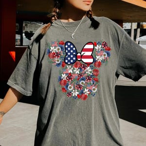 May include: Gray t-shirt featuring a floral design in the shape of a mouse head. The design includes a bow with an American flag pattern and is surrounded by red, white, and blue flowers and small American flags.