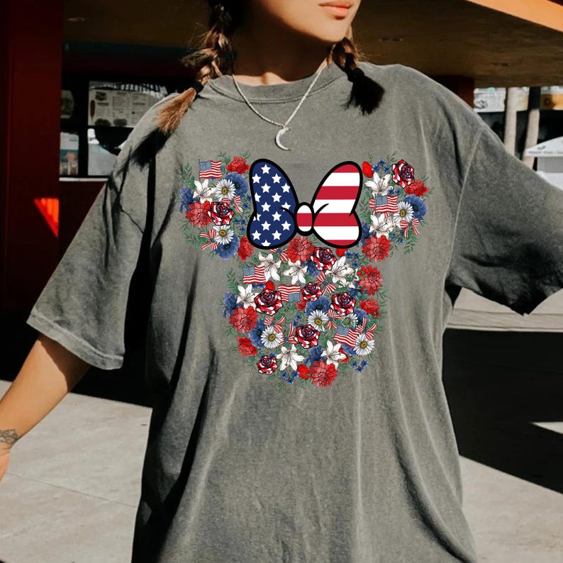 May include: Gray t-shirt featuring a floral design in the shape of a mouse head. The design includes a bow with an American flag pattern and is surrounded by red, white, and blue flowers and small American flags.