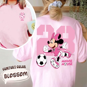 May include: Pale pink Comfort Colors t-shirt featuring a Minnie Mouse soccer design. The back displays a large pink number 23 and a Minnie Mouse illustration. The front has a small soccer graphic with the text "MINNIE".