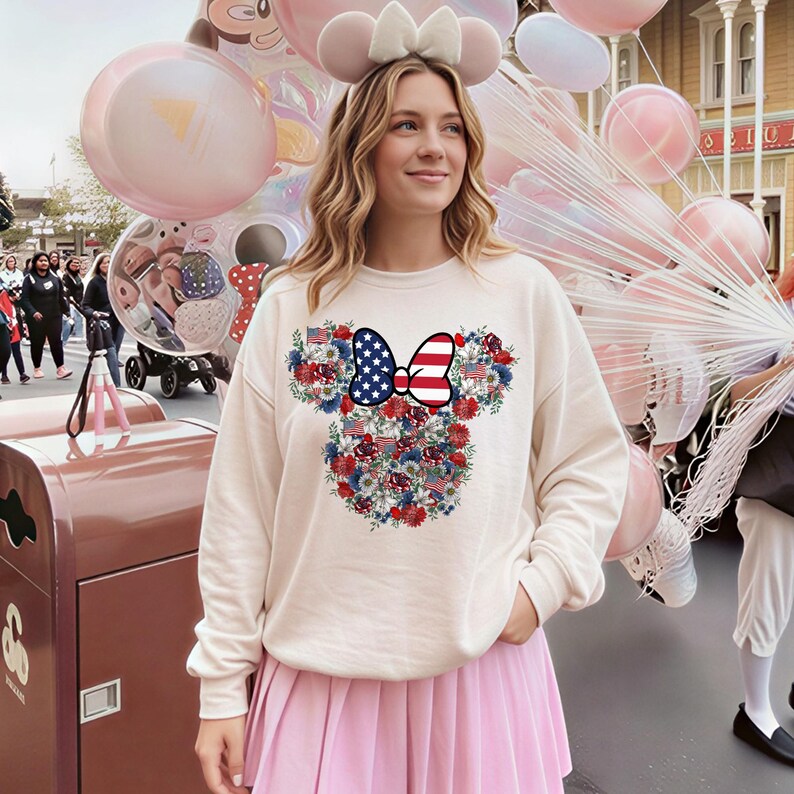 May include: Cream-colored sweatshirt featuring a Minnie Mouse design with a patriotic bow and floral accents. The bow is decorated with the American flag. The floral design includes red, white, and blue flowers and American flags.