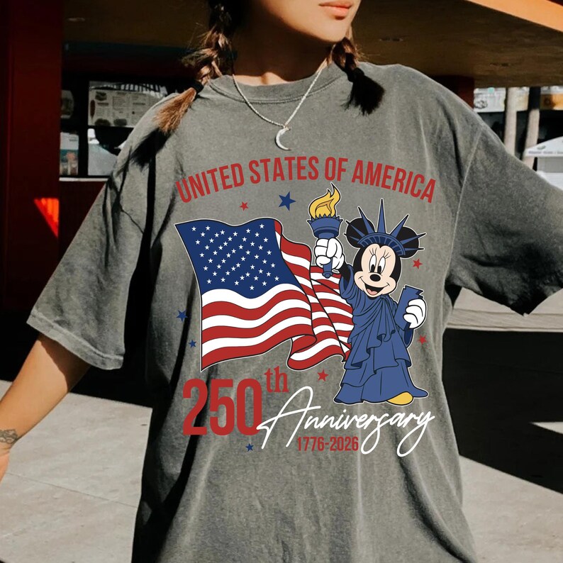 May include: Gray t-shirt featuring a graphic of the American flag, Mickey Mouse as the Statue of Liberty, and text that reads "United States of America 250th Anniversary 1776-2026". The design is in red, white, blue, and yellow.