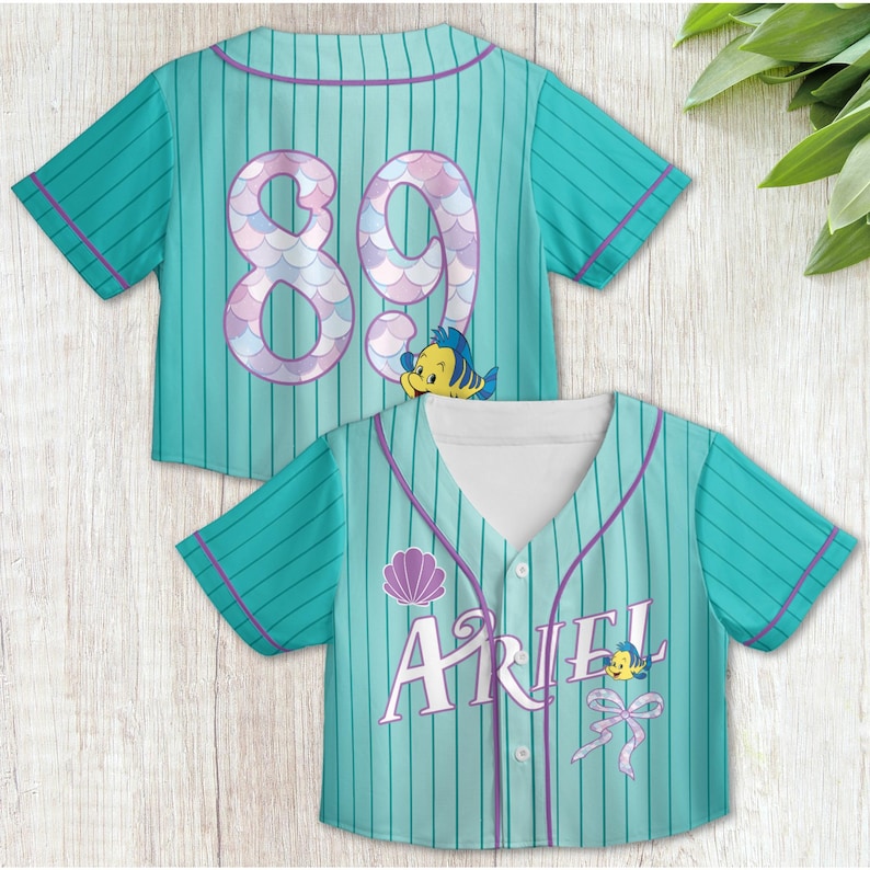 May include: A teal and purple baseball jersey with a mermaid-themed design. The back of the jersey has the number "89" in pink and purple with a Flounder character. The front of the jersey has the name "Ariel" in white with a seashell and Flounder character.