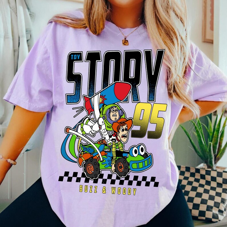 May include: A light purple t-shirt featuring a graphic of Buzz Lightyear and Woody from Toy Story in a race car. The text "TOY STORY 95" is above the graphic, with "Buzz & Woody" below.