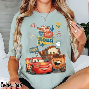 May include: Pale blue t-shirt with a graphic of Lightning McQueen and Mater from the film Cars, and the text "Let's Hit the Open Road". The design includes Route 66 signage and other road trip-themed elements.