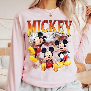 May include: A pale pink long-sleeved sweatshirt with a graphic of Mickey Mouse in three poses. The word "Mickey" is printed in red and yellow, with "Mouse" in cursive below.