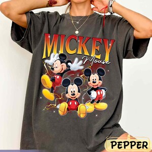May include: Dark grey t-shirt with a graphic of Mickey Mouse in three poses. The text "Mickey Mouse" is in red and yellow. The shirt has a vintage, distressed appearance.