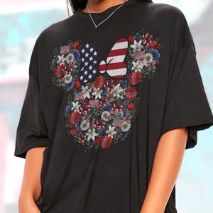 May include: Black t-shirt featuring a floral Minnie Mouse design with American flag elements. The design includes a bow with stars and stripes, surrounded by red, white, and blue flowers and small American flags.