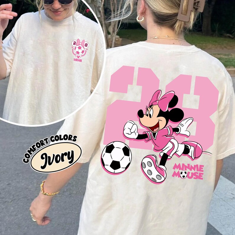 May include: Ivory t-shirt with a pink Minnie Mouse soccer design. The back shows a large pink number 23 with Minnie Mouse playing football. The shirt also has the words "Minnie Mouse" in pink.