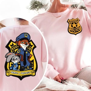 May include: Pale pink sweatshirt with a Zootropolis Police badge on the front and a larger graphic on the back. The graphic features Nick Wilde, Judy Hopps, and the words "Serve & Protect".