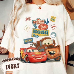 May include: Ivory t-shirt with a graphic of Lightning McQueen and Mater from the film Cars, with the text "Let's Hit the Open Road." The design includes Route 66 and road trip graphics.