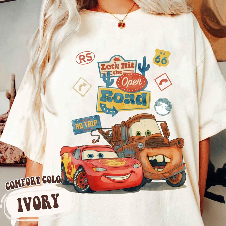 May include: Ivory t-shirt with a graphic of Lightning McQueen and Mater from the film Cars, with the text "Let's Hit the Open Road." The design includes Route 66 and road trip graphics.