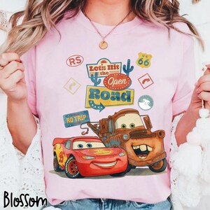 May include: A pale pink t-shirt with a graphic of Lightning McQueen and Mater from the film Cars. The design includes the text "Let's Hit the Open Road" and Route 66 signage. The word "Blossom" is printed at the bottom of the shirt.