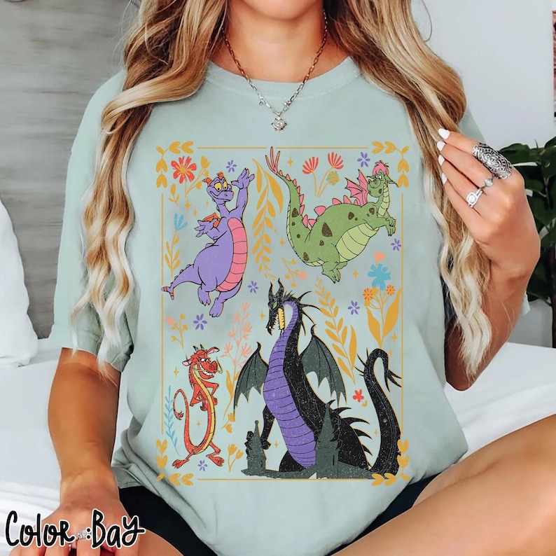 May include: Light blue t-shirt featuring a colorful graphic of cartoon dragons within a floral frame. The dragons are in various colors, including purple, green, red, and black. The text "Color Bay" is at the bottom left.