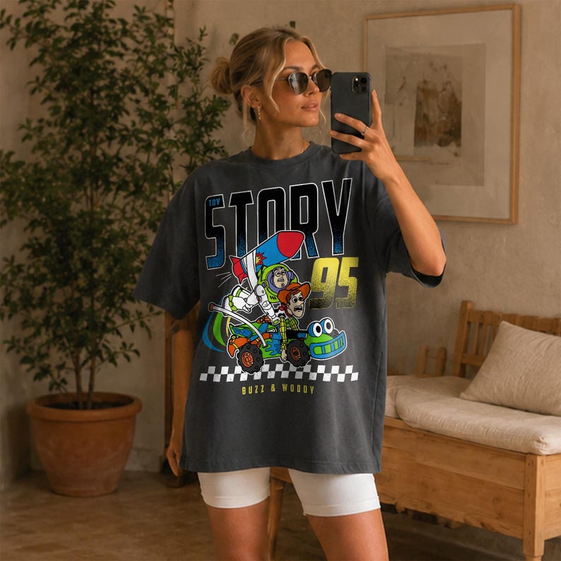 May include: Dark gray t-shirt featuring a graphic of Buzz Lightyear and Woody from Toy Story in a race car. The text "TOY STORY 95" is above the graphic, and "BUZZ & WOODY" is below. The shirt has a checkered flag design.