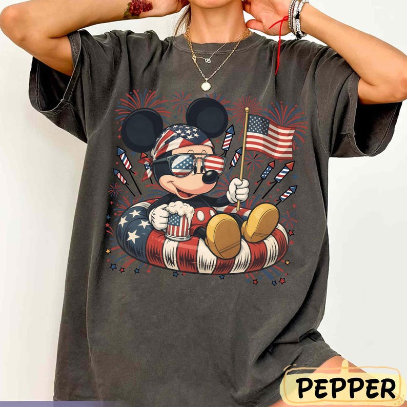 May include: Dark gray t-shirt featuring a patriotic Mickey Mouse design. Mickey is wearing sunglasses and a bandana with the American flag pattern, holding a beer mug and an American flag. The design includes fireworks and a red, white, and blue inner tube.
