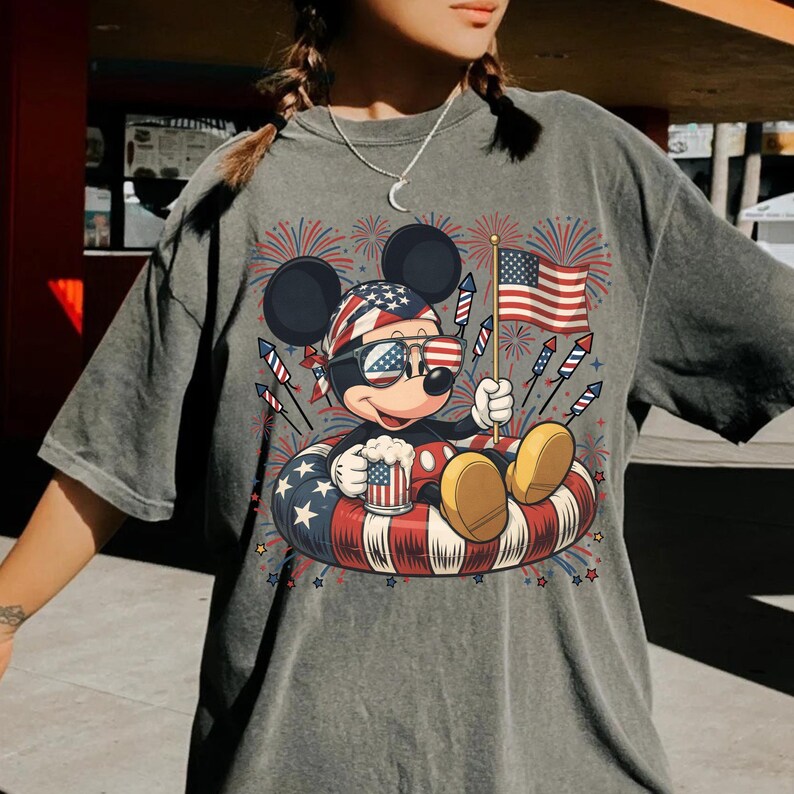 May include: Gray t-shirt featuring a patriotic Mickey Mouse design. Mickey is wearing sunglasses and a bandana with an American flag pattern, holding a beer mug and an American flag while floating on a red, white, and blue inner tube. Fireworks are in the background.