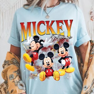 May include: Pale blue t-shirt with the words "Mickey Mouse" in a yellow and red gradient font. The shirt features three cartoon images of Mickey Mouse in different poses, wearing yellow shoes and red shorts.