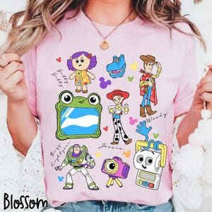 May include: A light pink t-shirt featuring colorful cartoon illustrations of characters from the Toy Story movie franchise. The shirt includes images of Woody, Buzz Lightyear, Jessie, and others, with the word "Blossom" at the bottom.