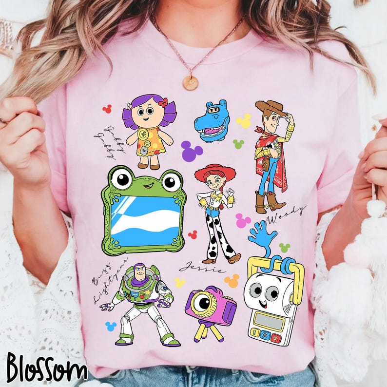 May include: A light pink t-shirt featuring colorful cartoon illustrations of characters from the Toy Story movie franchise. The shirt includes images of Woody, Buzz Lightyear, Jessie, and others, with the word "Blossom" at the bottom.