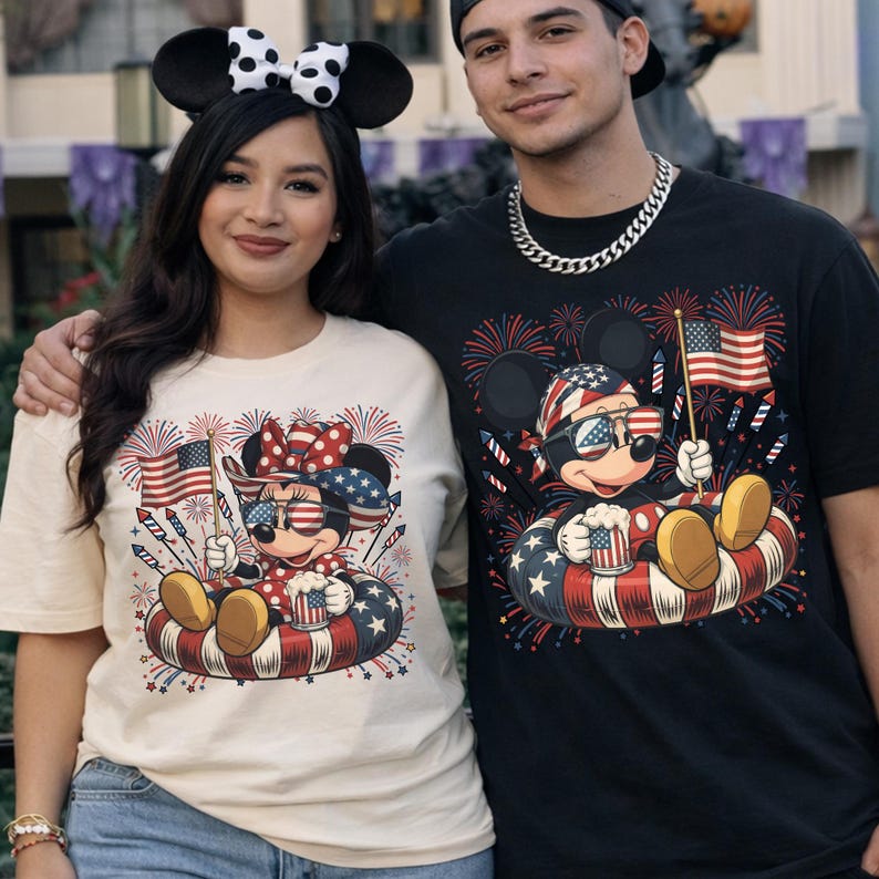 May include: Two people wearing matching graphic t-shirts featuring a patriotic Mickey Mouse design. The design includes fireworks, an American flag, and Mickey Mouse wearing sunglasses. One shirt is cream-colored, the other is black.