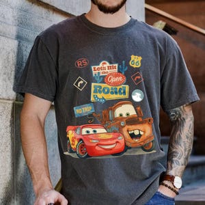 May include: Dark grey t-shirt featuring Lightning McQueen and Mater from the film Cars. The shirt has a graphic with the text "Let's Hit the Open Road" and Route 66 signage.