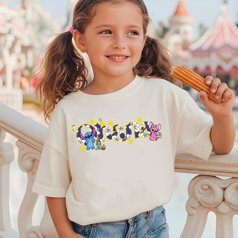 Puede incluir: Camiseta color crema con la palabra "Ohana" en un dise&ntilde;o floral con los personajes de Stitch y Angel. La camiseta es de corte holgado y la lleva una ni&ntilde;a.