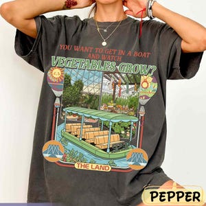 May include: Dark grey t-shirt featuring the text "YOU WANT TO GET IN A BOAT AND WATCH VEGETABLES GROW?" and a graphic of a boat ride through a greenhouse. The design includes hot air balloons and the words "THE LAND".