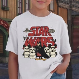 Puede incluir: Camiseta blanca con el texto rojo "STAR WARS" con efecto desgastado. El diseño incluye ilustraciones de naves de Star Wars, Darth Vader y soldados de asalto con temática de Mickey Mouse. Una camiseta gráfica divertida.