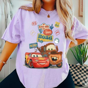 May include: A lilac t-shirt with a graphic of the Cars characters Lightning McQueen and Mater. The design includes the text "Let's Hit the Open Road" and other road trip-themed elements.