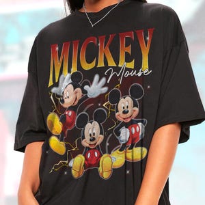 May include: Black t-shirt with a graphic of Mickey Mouse in different poses. The word "MICKEY" is in large, red and yellow letters above "Mouse" in a cursive font. The design includes lightning bolt accents.