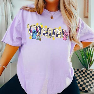 Puede incluir: Una camiseta de color lila claro con la palabra "Ohana" en un dise&ntilde;o floral amarillo y blanco. La camiseta presenta los personajes de dibujos animados Stitch, Angel y una rana. La camiseta es de manga corta.
