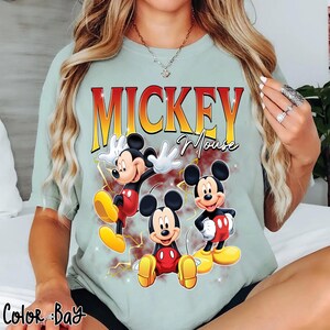 May include: Mint green t-shirt featuring three Mickey Mouse illustrations with the text "MICKEY Mouse" in a gradient orange and yellow font. The cartoon characters are in red, black, white, and yellow. The shirt has a relaxed fit.