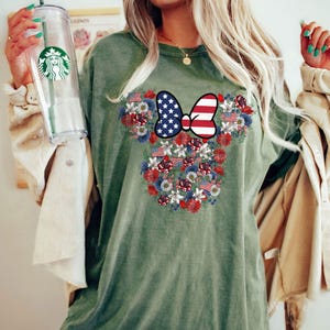 May include: An olive green t-shirt featuring a floral design in the shape of a mouse head. The design incorporates red, white, and blue flowers, American flags, and a bow with a stars and stripes pattern.