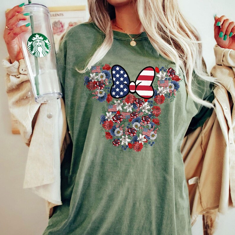 May include: An olive green t-shirt featuring a floral design in the shape of a mouse head. The design incorporates red, white, and blue flowers, American flags, and a bow with a stars and stripes pattern.