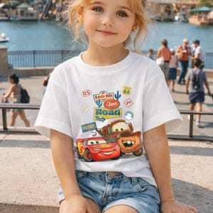 May include: White t-shirt with a graphic of Lightning McQueen and Mater from the film Cars, and the text "Lets Hit the Open Road". The shirt is worn by a young girl, paired with denim shorts.